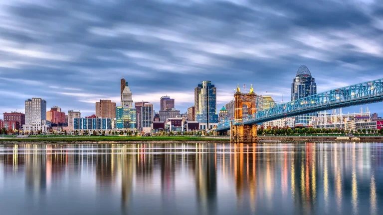 Cincinnati skyline across the Ohio River at dusk, with a lit blue steel suspension bridge on stone towers at right and the buildings reflected in the water under streaked clouds