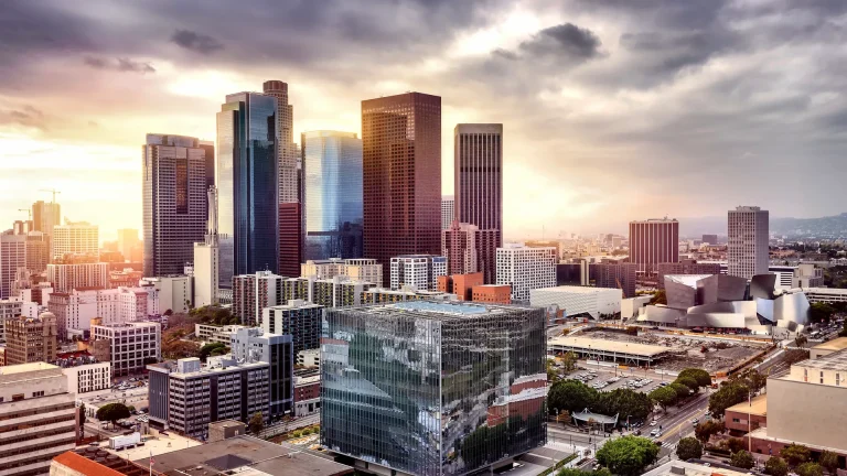 Downtown Los Angeles seen from above in warm evening light, with glass and brick office towers, a glass-clad cube-shaped building in the foreground and a curved silver steel-clad building to the right under a cloudy golden sky