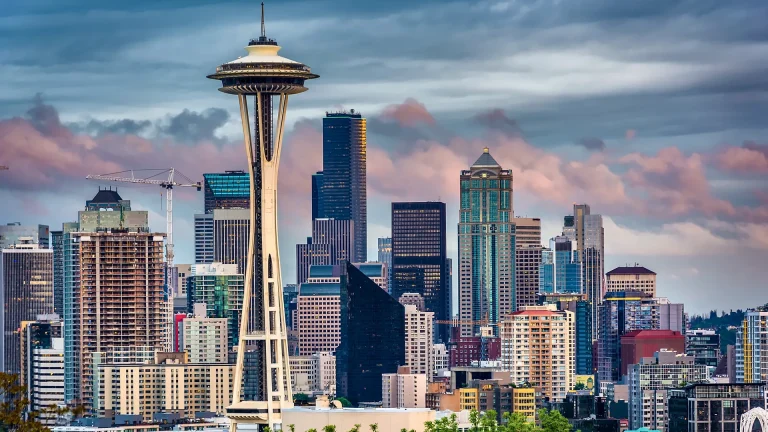 The Space Needle rises in front of downtown Seattle's towers under pink-tinged evening clouds, with a construction crane at the left edge