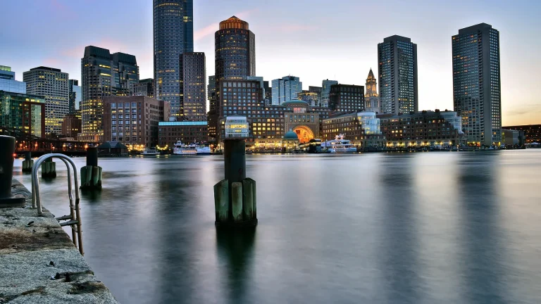 Boston's waterfront at dusk, with lit office towers and a clock tower across calm harbor water, a granite quay on the left and a wooden piling in the foreground