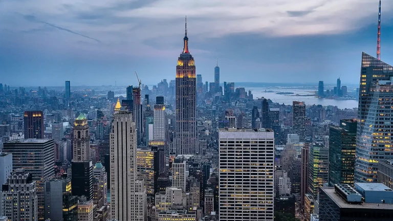 Midtown Manhattan at dusk, with the Empire State Building lit near its top, dense office towers on every side and the lower Manhattan skyline and water beyond under a cloudy sky