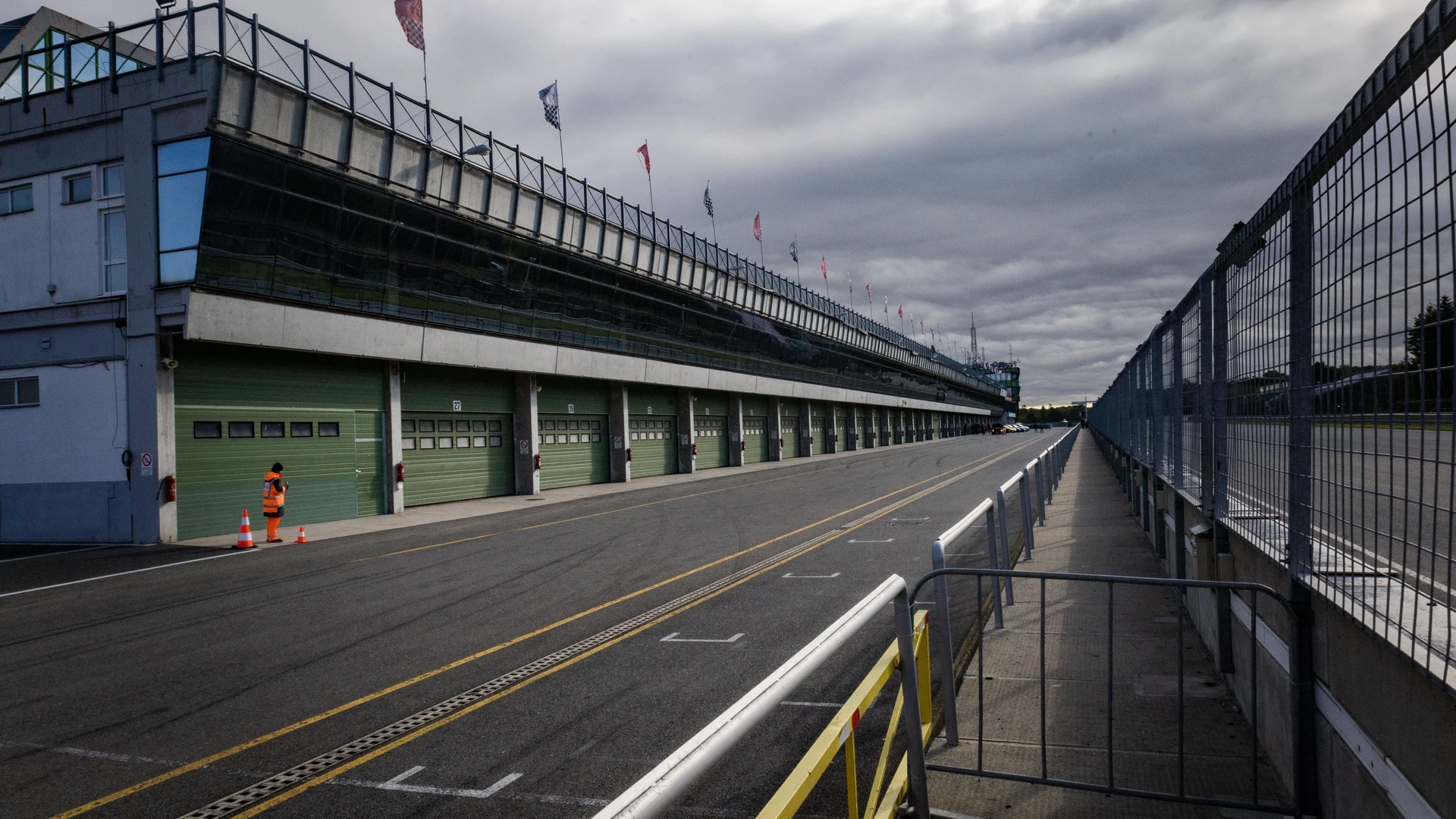 An empty pit lane at a motor racing circuit, with a long row of green garage doors under a flag-topped building on the left, a fenced walkway on the right and one person in an orange vest standing near traffic cones under an overcast sky