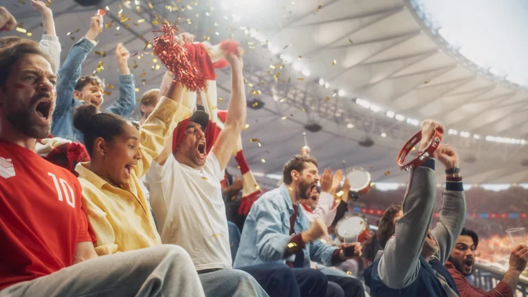 Fans in red and white shirts shout and cheer in the stands under a stadium roof, raising fists, a red pom-pom, a scarf and a tambourine while gold confetti falls
