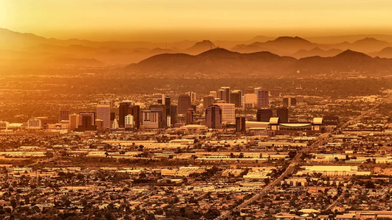 Downtown Phoenix towers above a wide grid of low-rise neighborhoods, with layered mountain ridges behind them in warm golden haze