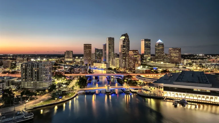 Downtown Tampa at dusk, with lit office towers rising beyond a river crossed by two illuminated bridges, and a low, brightly lit waterfront building with docked boats at the right, under an orange and blue sky