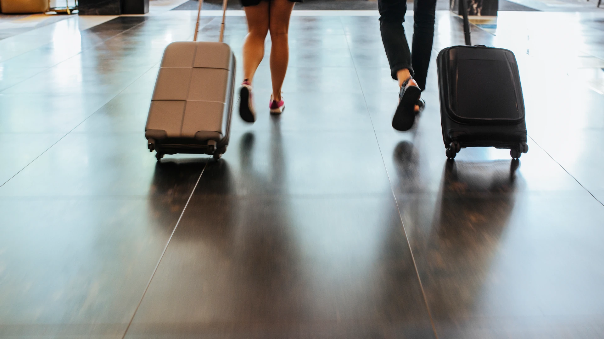 Two travelers walk away from the camera pulling rolling suitcases across a glossy floor